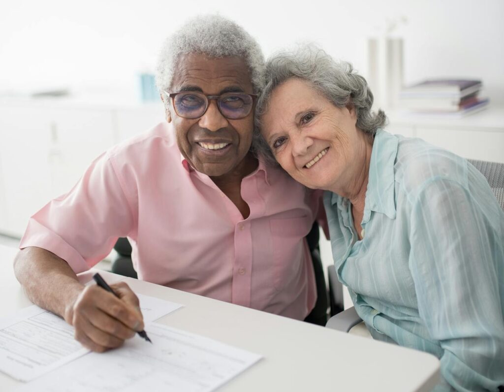 A retiree couple is smiling at the camera while signing the closing documents for their portfolio loan. 
